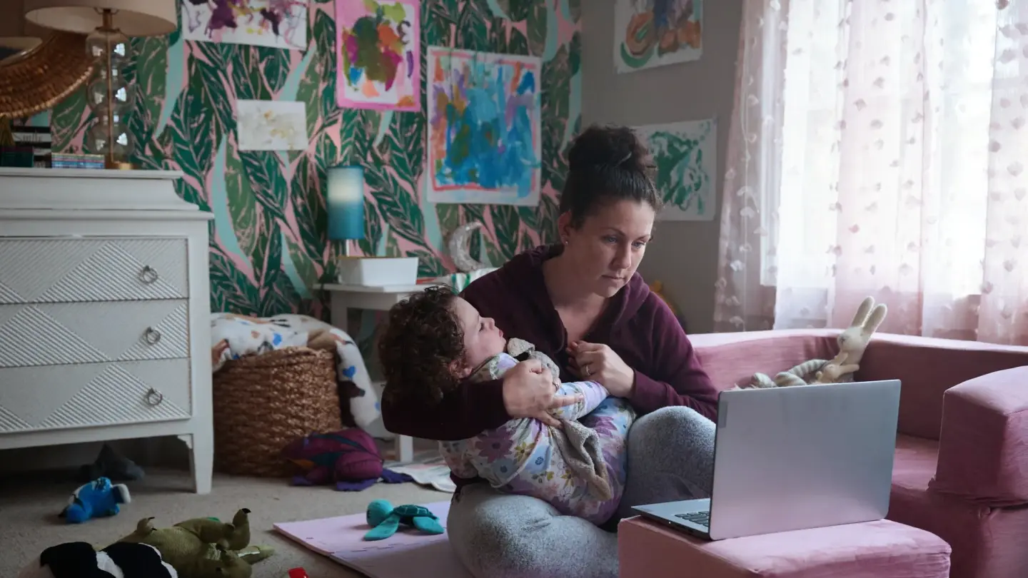A mother holding her young child while on a video call on her computer. They are sitting in the middle of a playroom in their house.