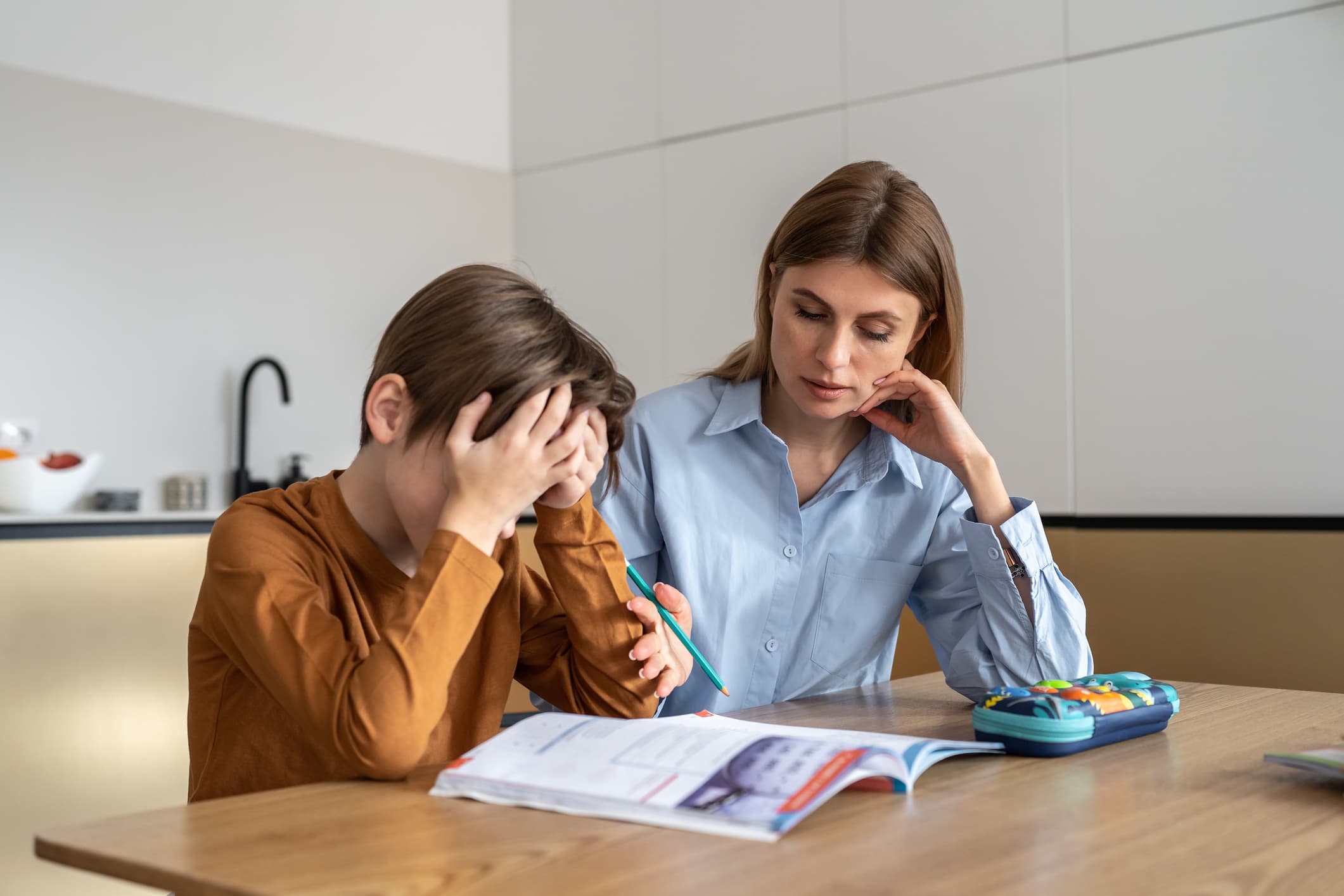 Boy seated at a table feeling frustrated while doing homework with his mother at home.
