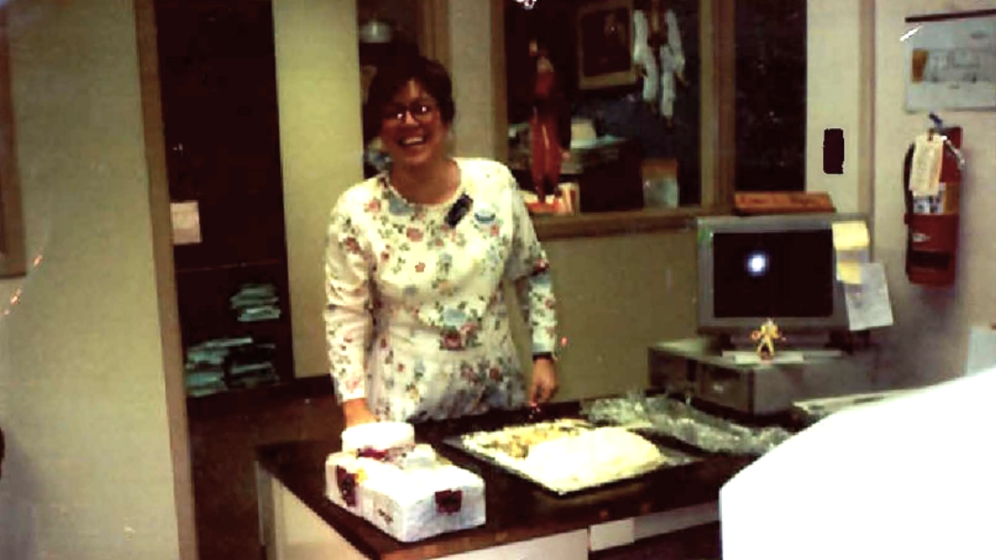 Betty Nuñez stands in a hospital hallway, smiling while using a rolling workstation with folders and a computer on top.