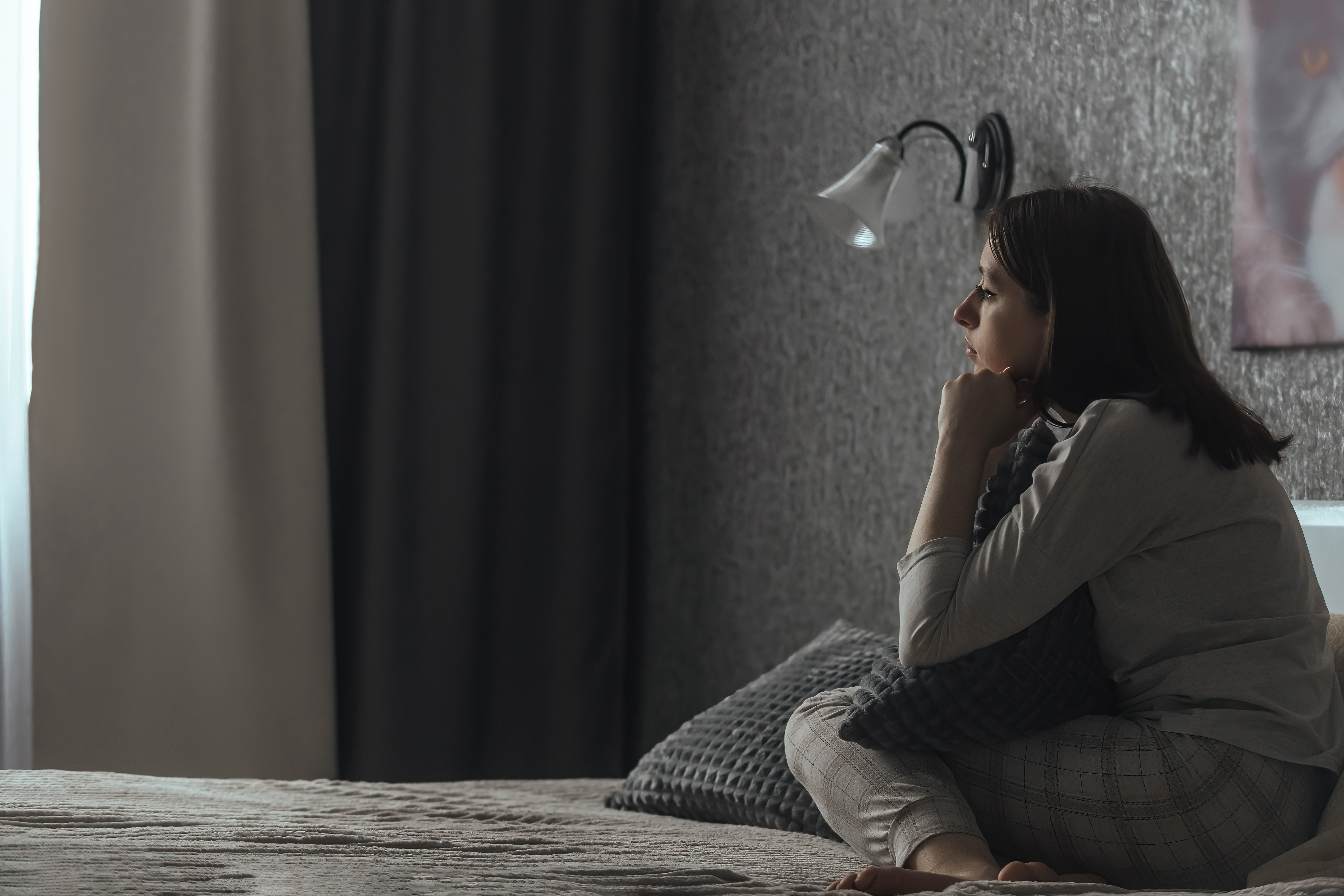 A sad young woman in pajamas sits alone on the bed and looks out the window.