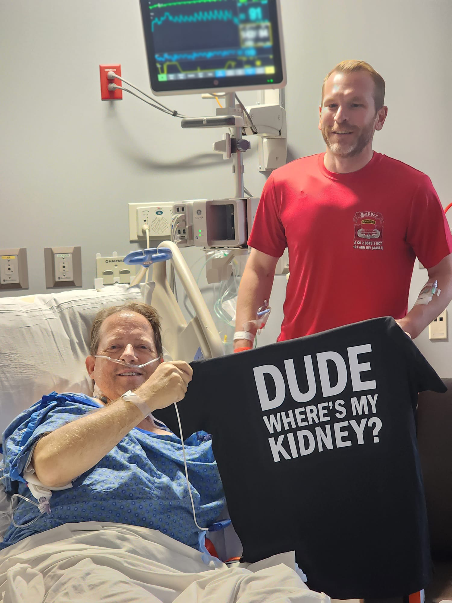 Man in hospital bed with coworker standing beside him, holding black shirt that says "dude, where's my kidney?"