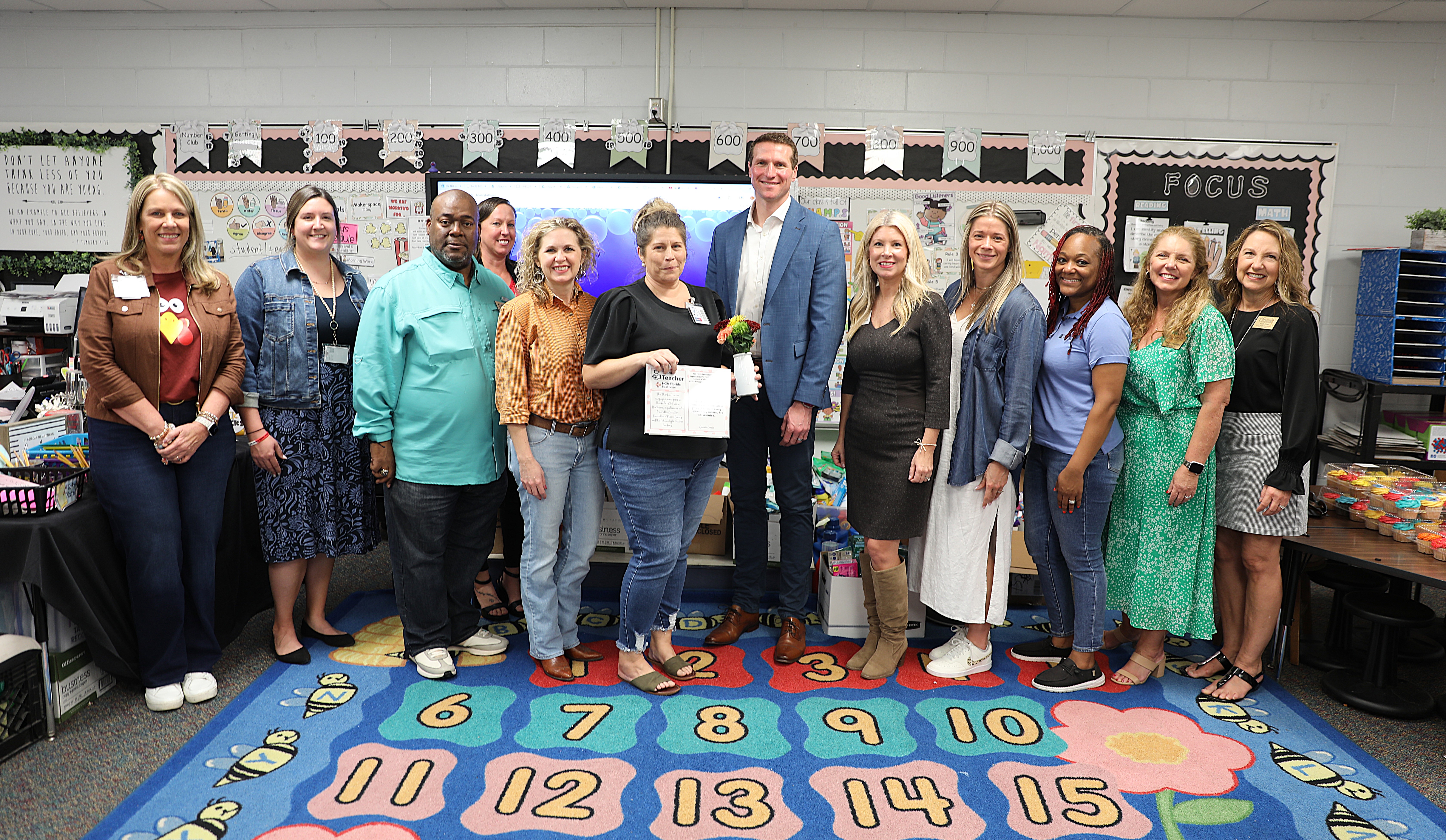A group of 10 women and 2 men stand next to each other, smiling while the woman in the middle holds a certificate and a small bouquet of flowers.