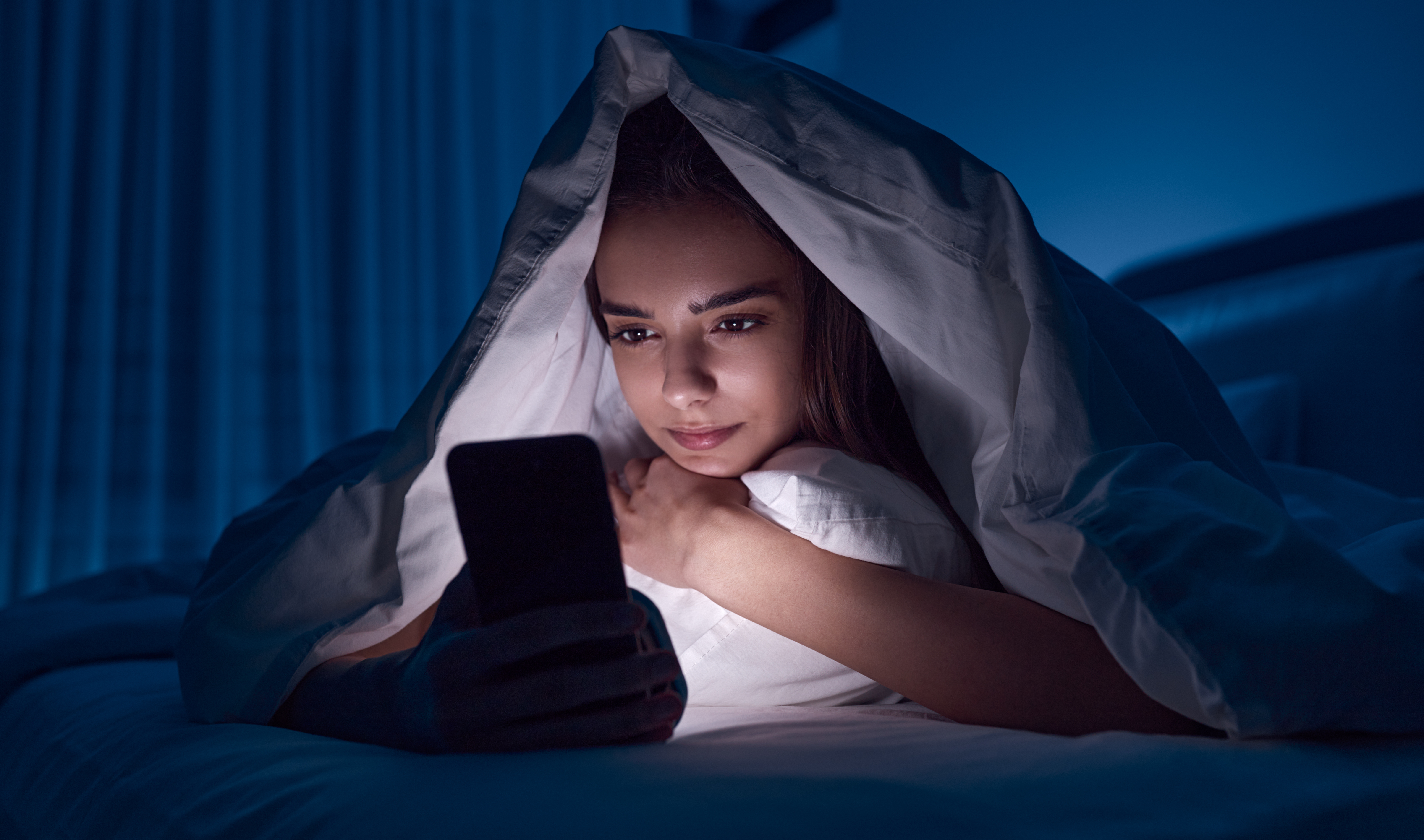 Young woman covered with a blanket in bed browsing on a mobile phone at night before going to sleep.