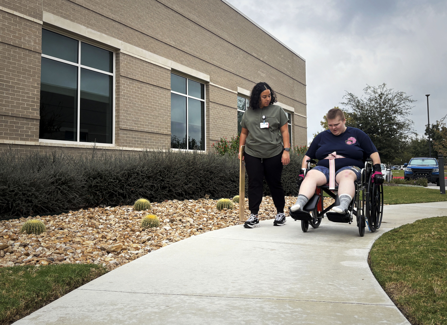 A person in a wheelchair moves along a curved sidewalk outside a modern brick building, accompanied by a staff member walking beside them. The staff member holds a wooden cane, and the landscaped area next to the sidewalk features rocks and small plants.