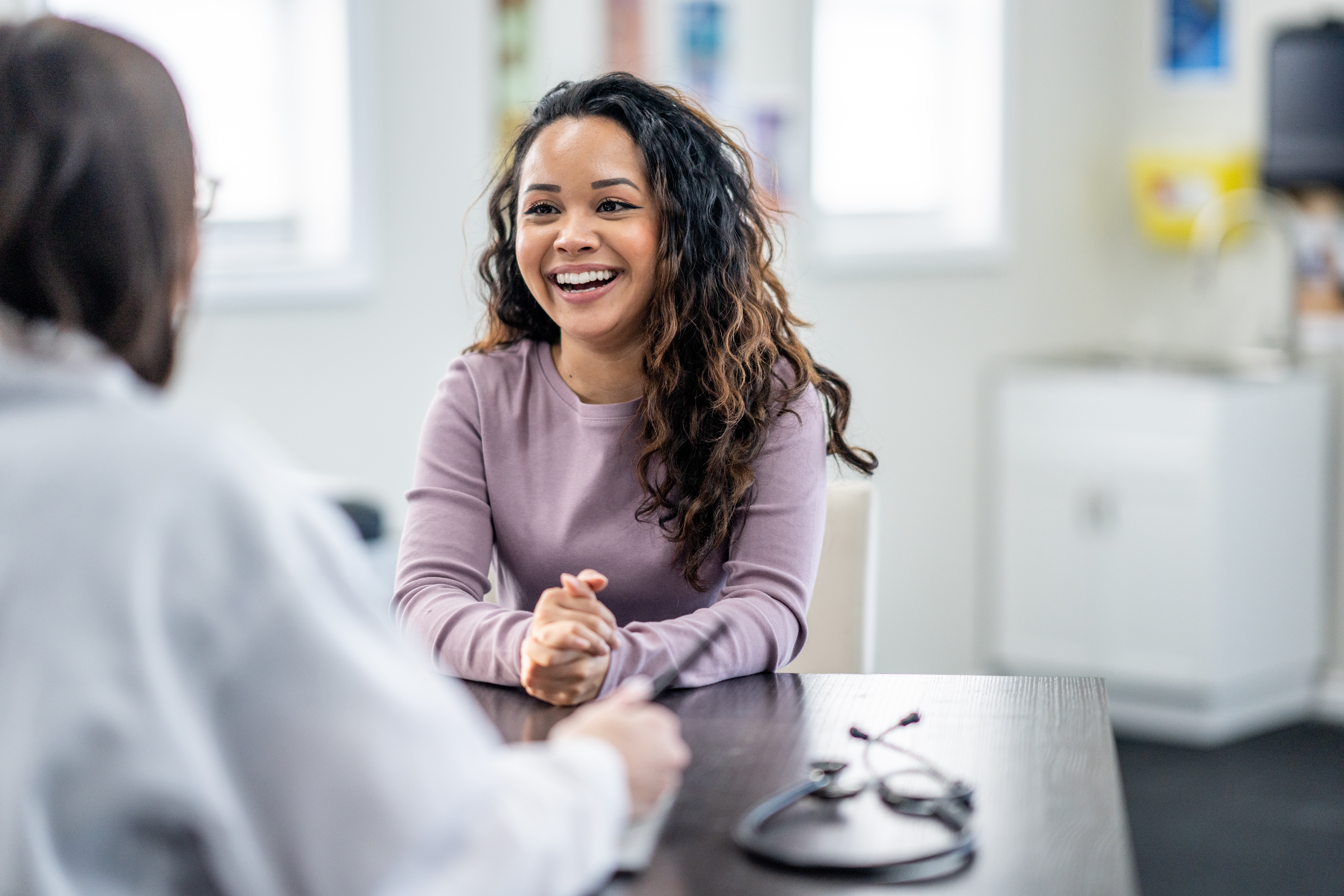 A smiling young patient sits with her doctor in an examination room, discussing her health concerns.