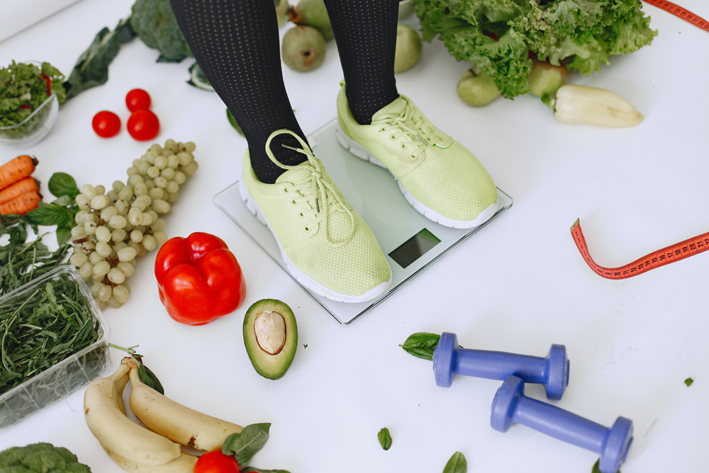 A person standing on a scale, surrounded by fresh fruit and vegetables, hand weights, and measuring tape.