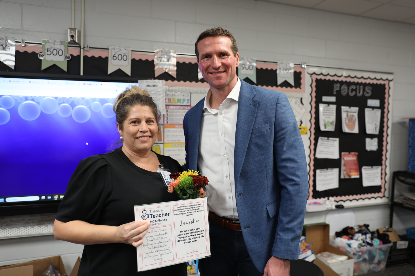 Shows a man in a blue suit and a woman standing next to each other smiling, as the woman holds a certificate and a small bouquet of flowers.