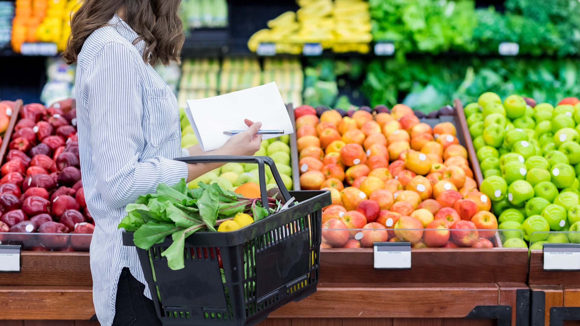 A woman holding a basket filled with fresh vegetables walks past the fruit display at a supermarket.
