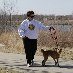 Joan walks with her dog on a sidewalk near a nature area.