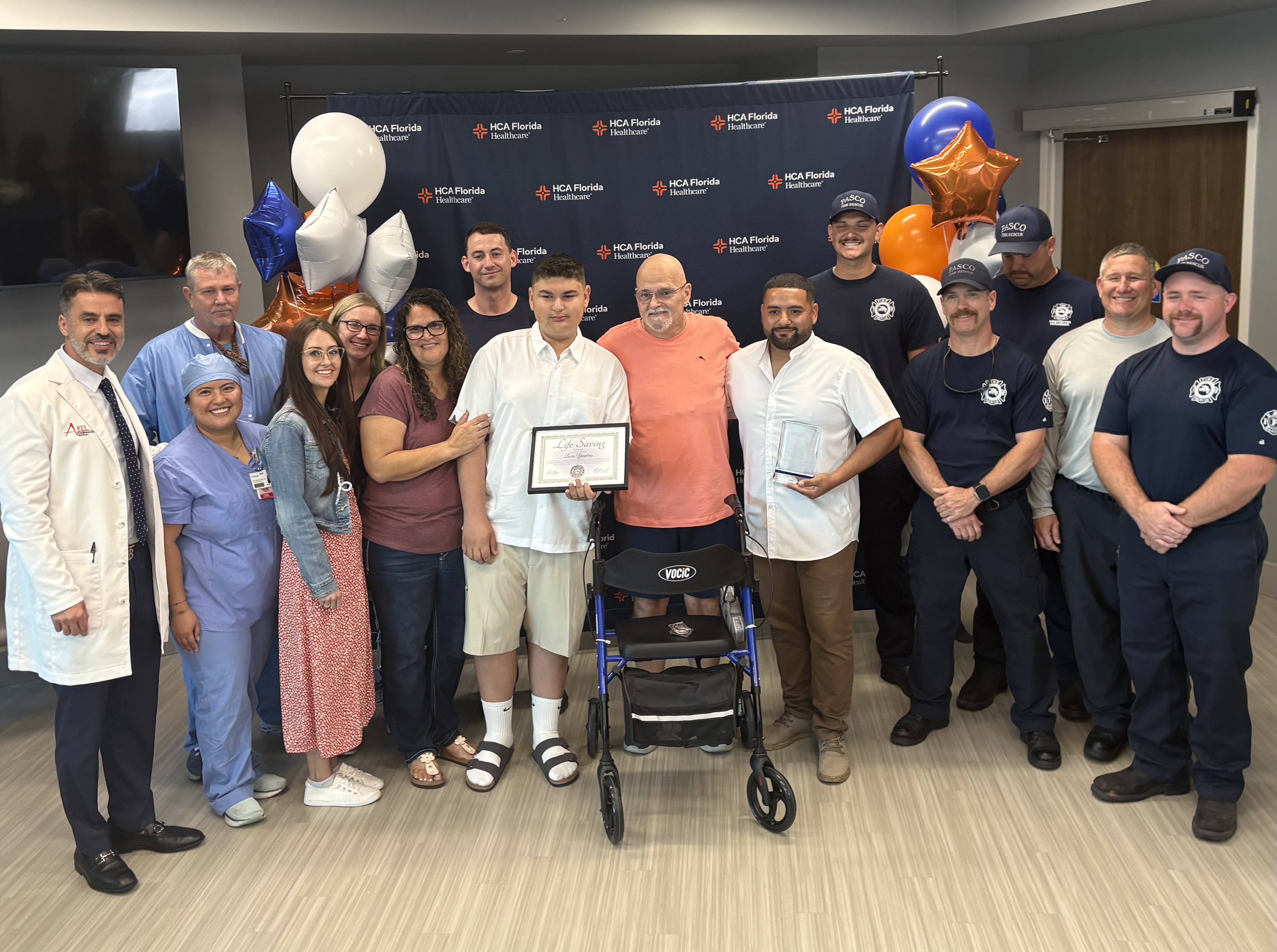Group photo of Randy Yingling in front of a walker/wheelchair surrounded by hospital care team members, first responders, and volunteers involved in his care. The group is standing indoors in front of an HCA Florida Healthcare backdrop with blue, orange, and white balloons, posing together during a recognition celebration.