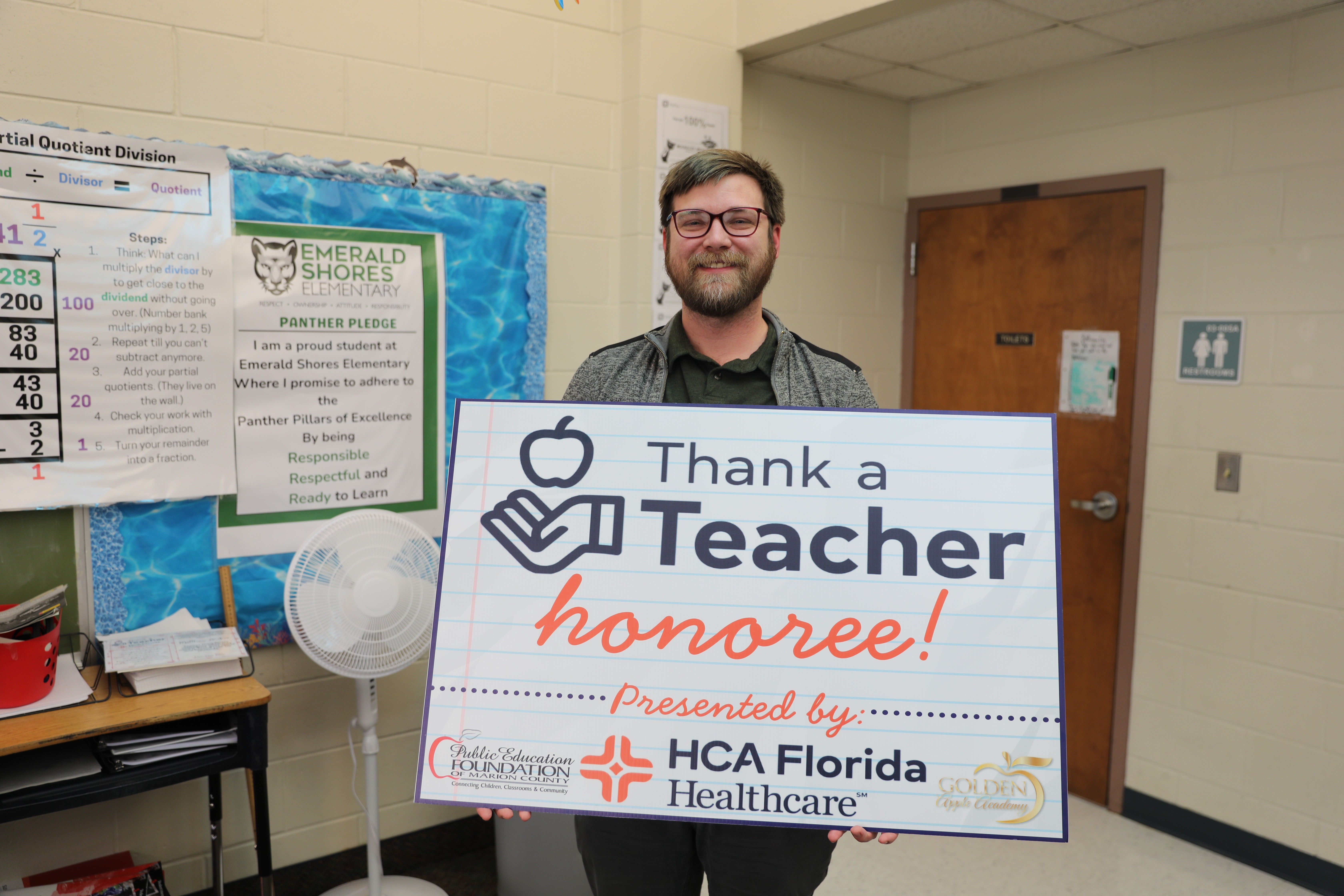 Matthew Andres stands in a classroom holding a "Thank a Teacher honoree" sign presented by HCA Florida Healthcare and the Public Education Foundation of Marion County, with school posters visible behind him.