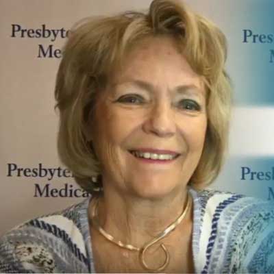 Nancy Moeglein smiles while wearing a colorful pattered blouse and is standing in front of a banner with the Presbyterian/St. Luke's Medical Center logo.