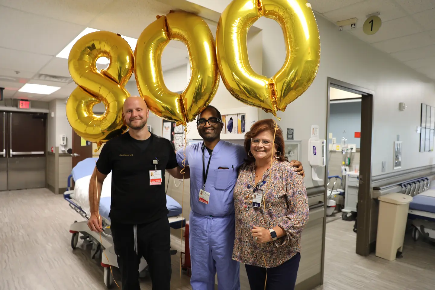 3 people standing in hospital hallway with gold balloons that spell out "800"