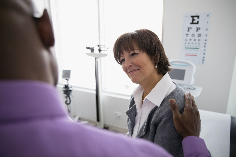 A person sitting on an exam table speaks with a healthcare professional who has a hand placed supportively on their shoulder.