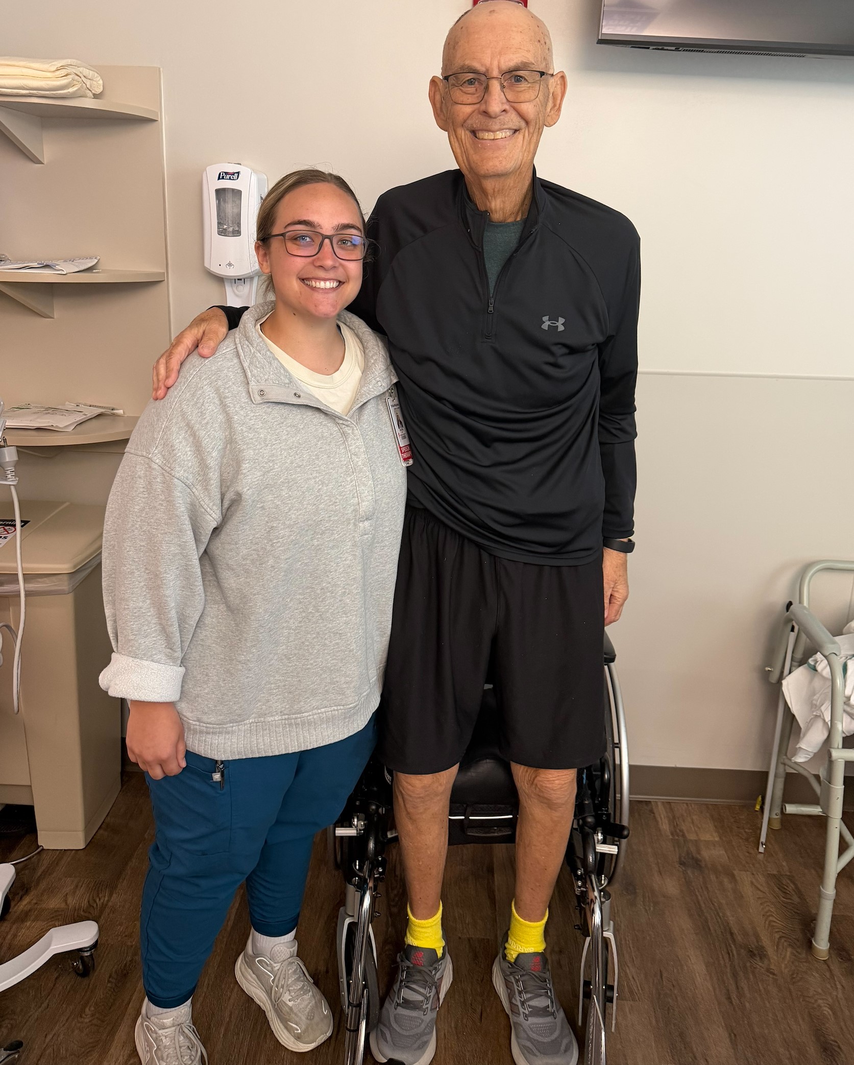 A smiling healthcare professional wearing glasses, a light gray quarter-zip sweatshirt, teal scrub pants, and white sneakers stands beside a smiling older adult wearing glasses, a black long-sleeve athletic shirt, black shorts, gray athletic shoes, and bright yellow socks. The older adult, Terry, stands in front of a wheelchair with one arm around the healthcare professional's shoulder. They are posing together in a rehabilitation clinic with a walker, shelving, and a wall-mounted hand sanitizer dispenser visible in the background.