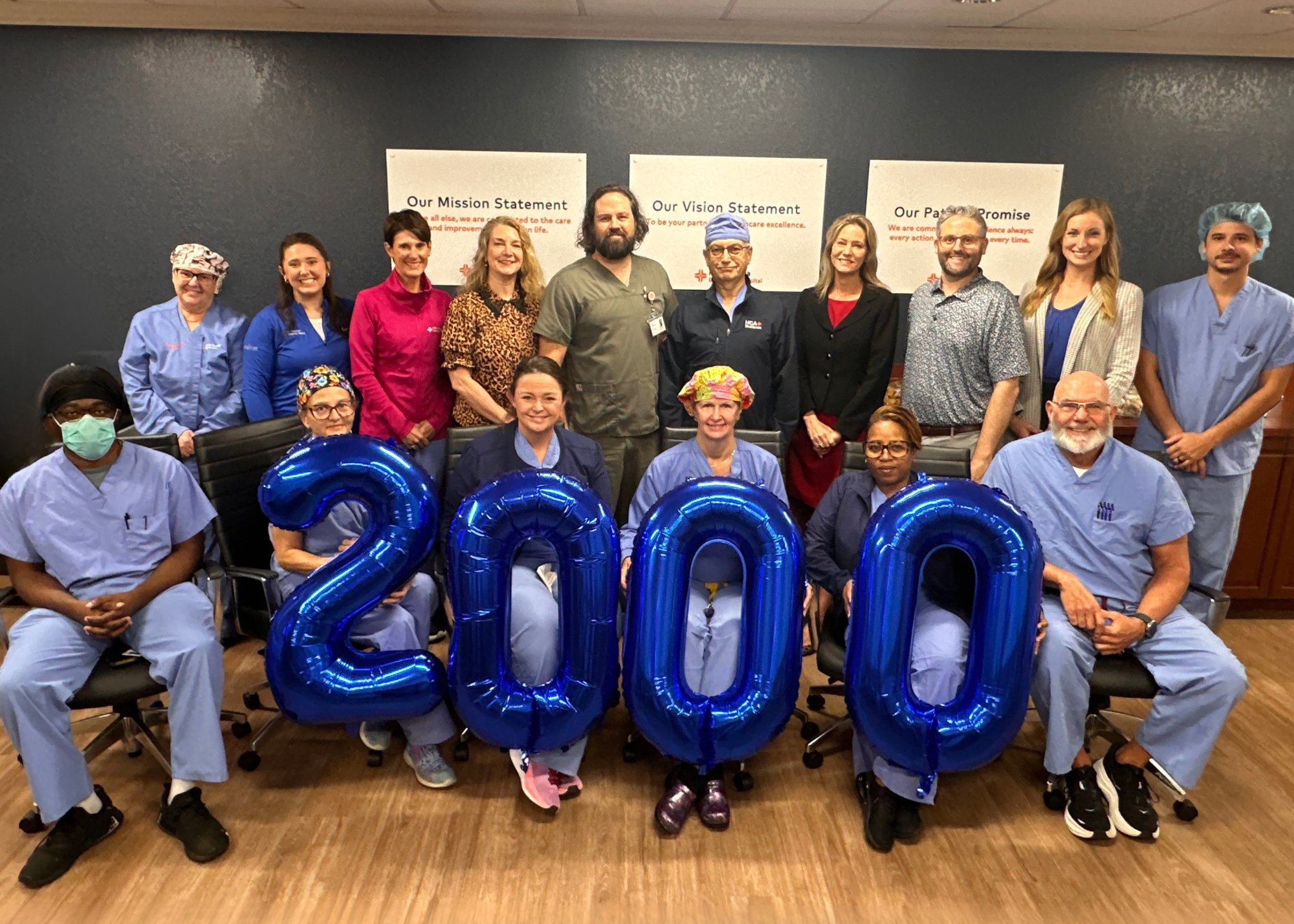 A group of hospital surgery team members pose indoors in front of posters displaying the mission, vision, and promise statements. Several people seated in the front row hold large blue balloon numbers forming "2000," celebrating the milestone of 2,000 robotic surgeries. The group is dressed in a mix of scrubs and professional attire.