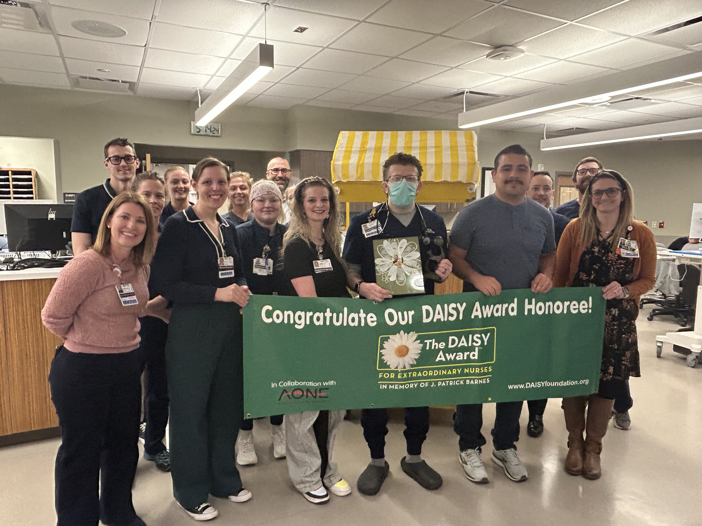 Group of healthcare staff standing together in a hospital unit, holding a large green banner that reads ‘Congratulate Our DAISY Award Honoree!’ with the DAISY Award logo.