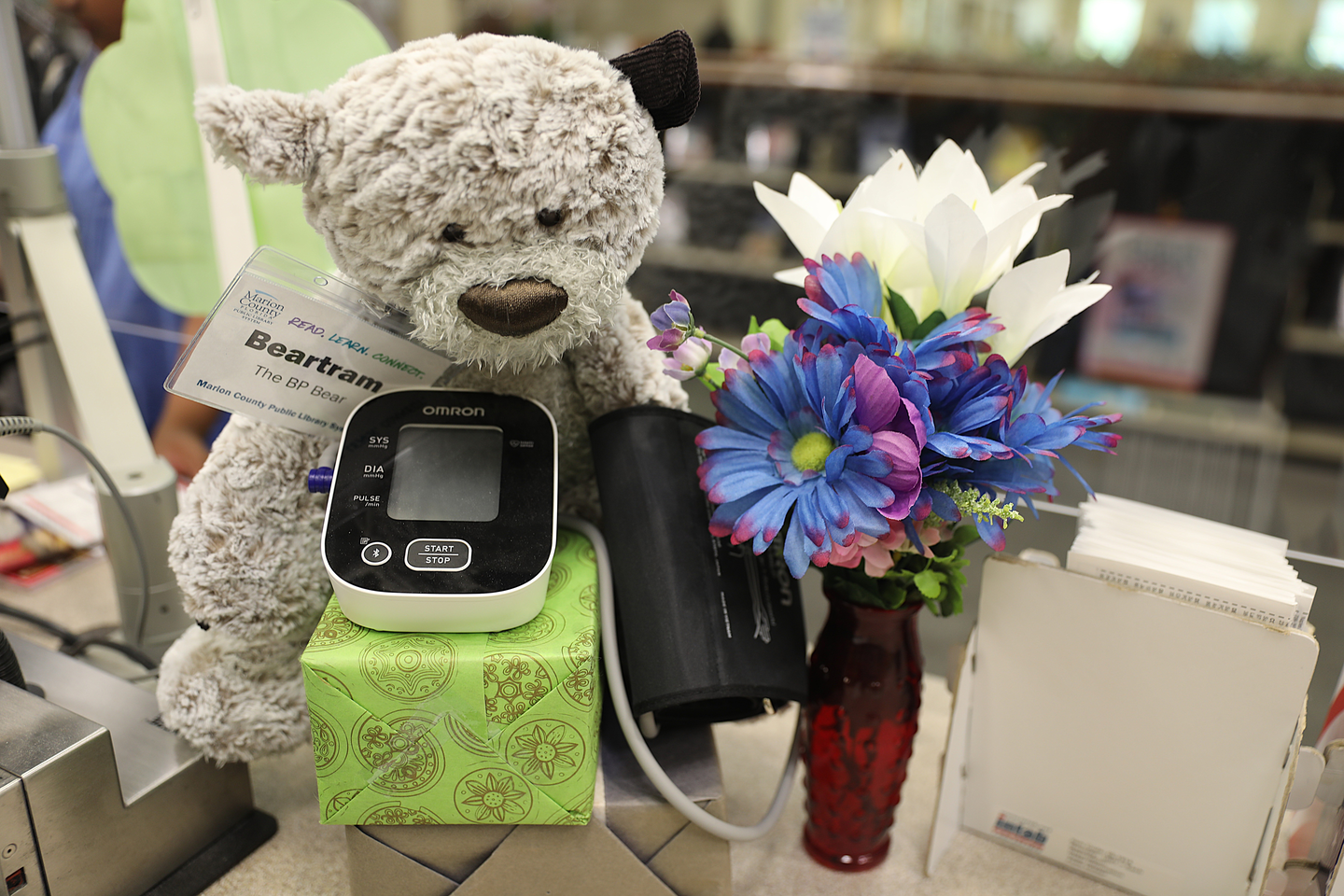 A gray teddy bear wearing a Marion County Public Library staff name badge reading "Beartram" sits at a public service desk beside an Omron blood pressure monitor. A colorful vase of blue, purple, and white flowers and a file holder are nearby, highlighting the library's health and wellness resources.