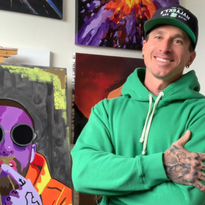 A man in a green hoodie stands in front of colorful paintings and smiles at the camera.