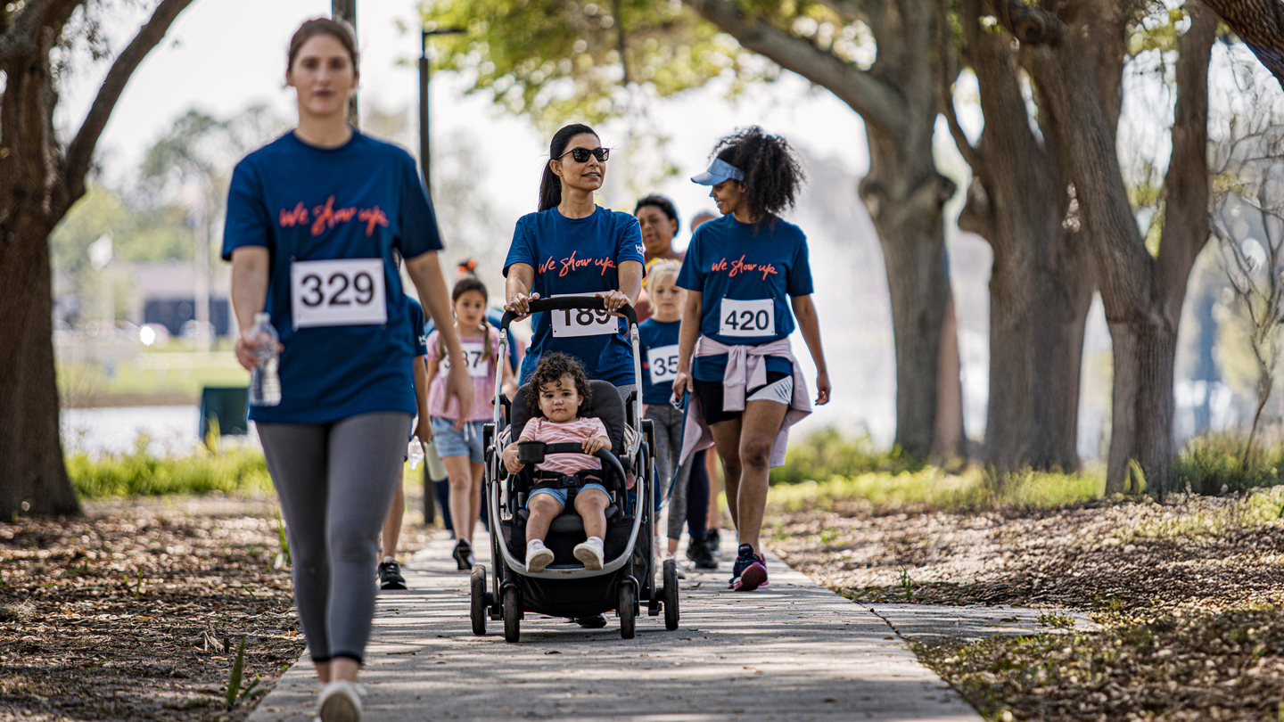 A group of women and children walking on the sidewalk wearing shirts that say We Show Up® and wearing race bibs.