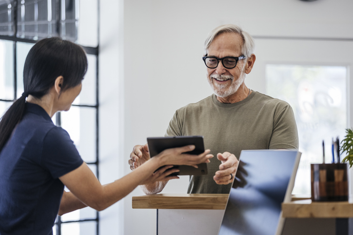 Older adult male in olive green shirt filling out a form on tablet, at reception desk with clinic worker.