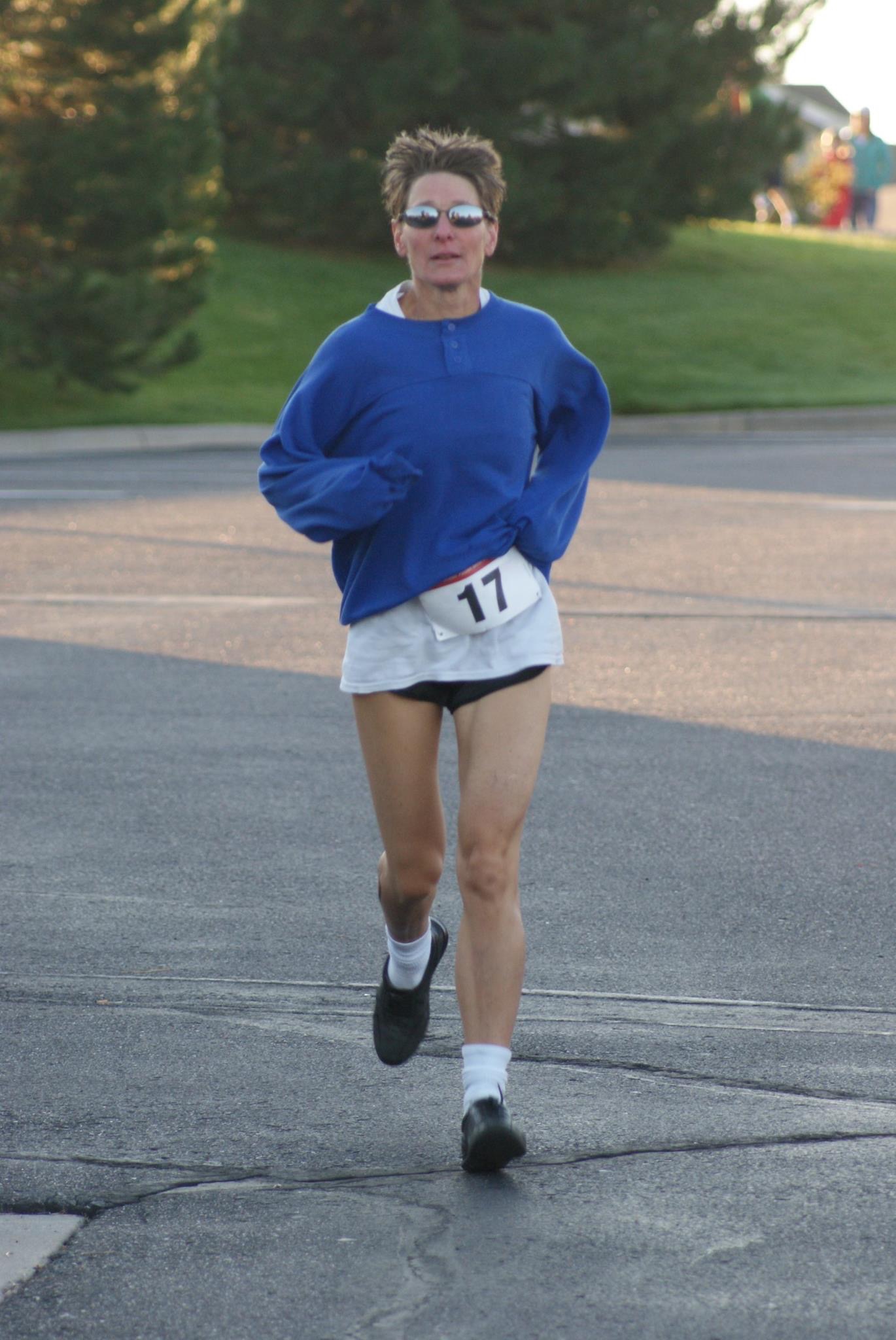 Sandra Kelly wearing a blue long‑sleeve top, white shorts with the number 17 pinned on them, and black running shoes is jogging on a paved road during daylight, with grass and trees visible in the background. 