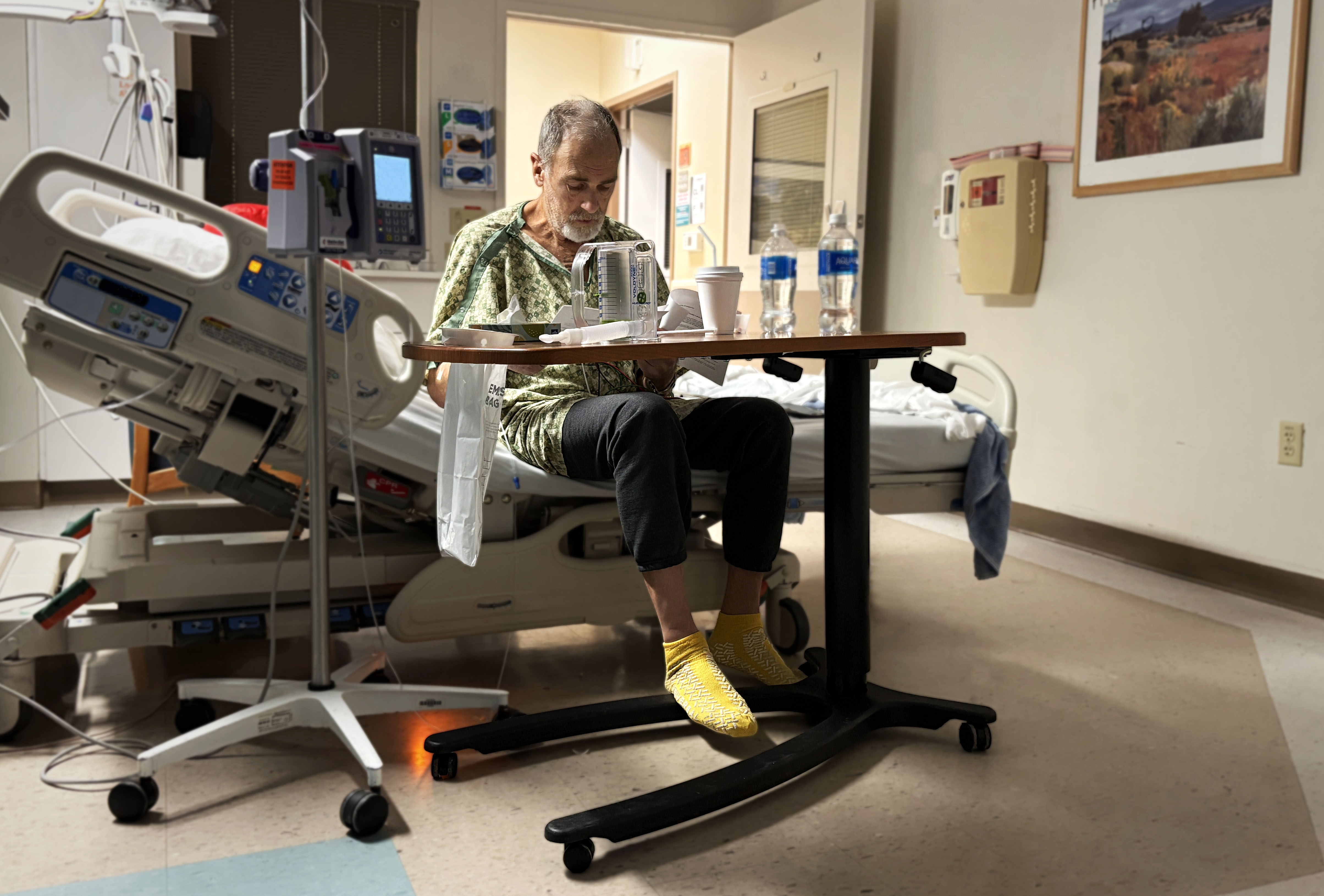 Harold Lance sits on a hospital bed in a patient room while reading papers at a bedside table after heart surgery.