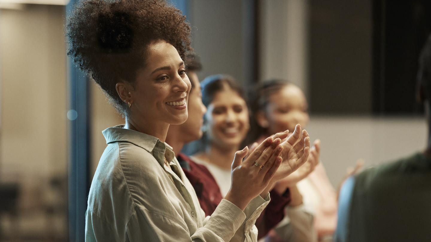 A woman in a workplace meeting with a team of coworkers, clapping in celebration.