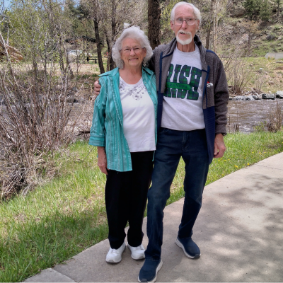 Older couple standing arm in arm on a path beside a creek in a wooded park.