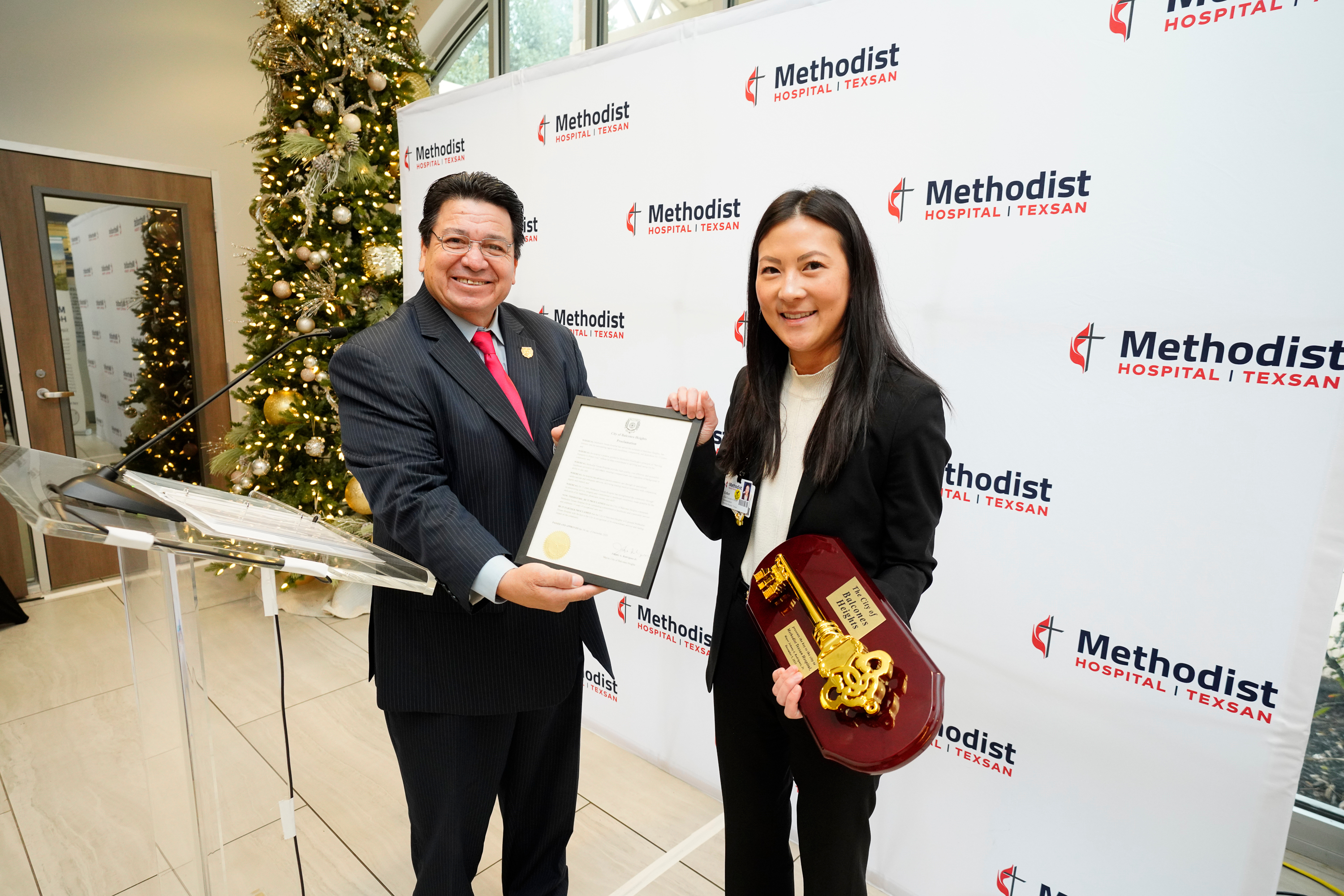 Two individuals stand in front of a Methodist Hospital Texsan backdrop. One person is holding a framed proclamation while the other holds a ceremonial oversized key, with a decorated Christmas tree partially visible behind them.