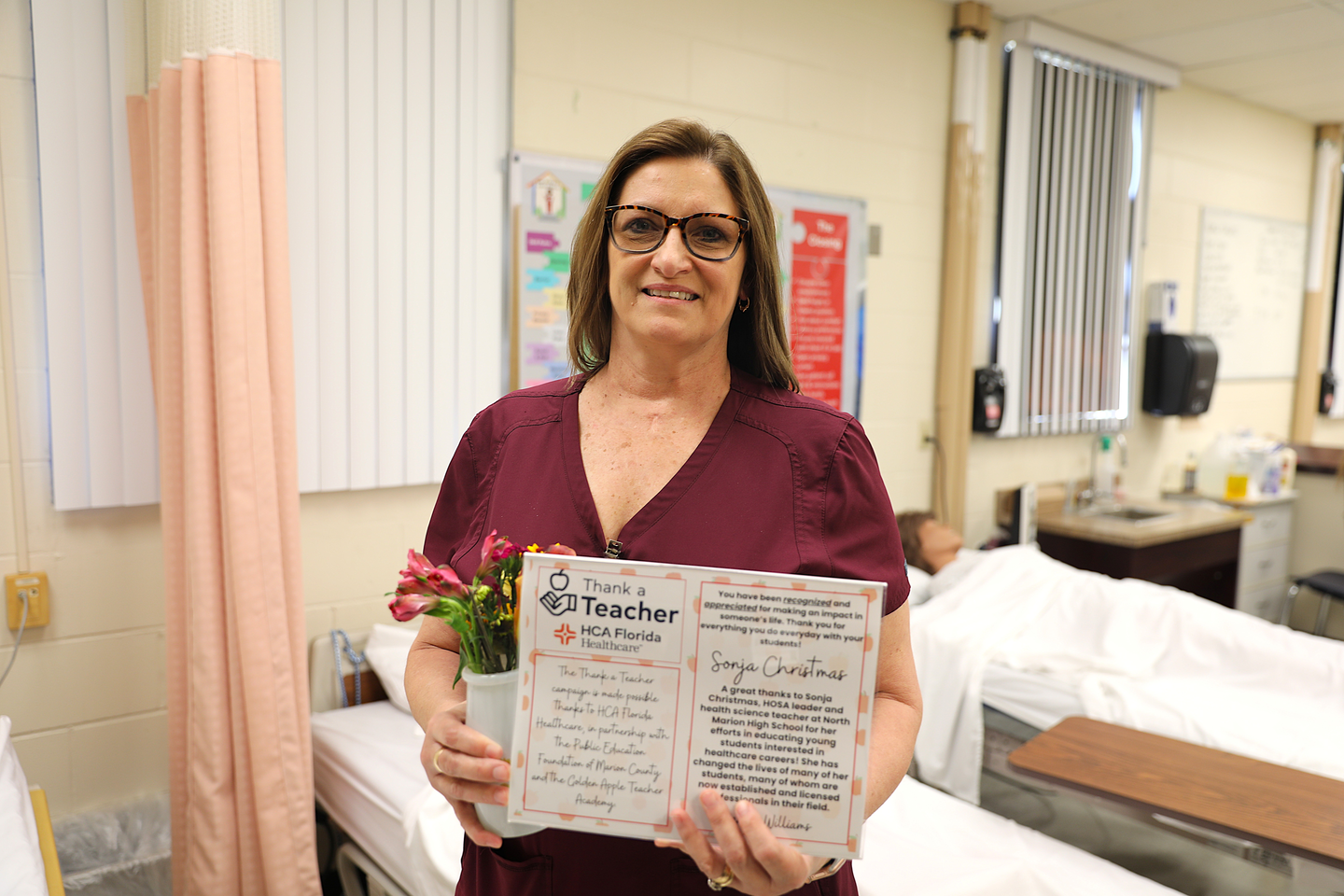 A woman wearing maroon medical scrubs stands in a classroom healthcare lab, holding a small bouquet of flowers and an open certificate that reads "Thank a Teacher – HCA Florida Healthcare." A hospital bed and medical training equipment are visible in the background.