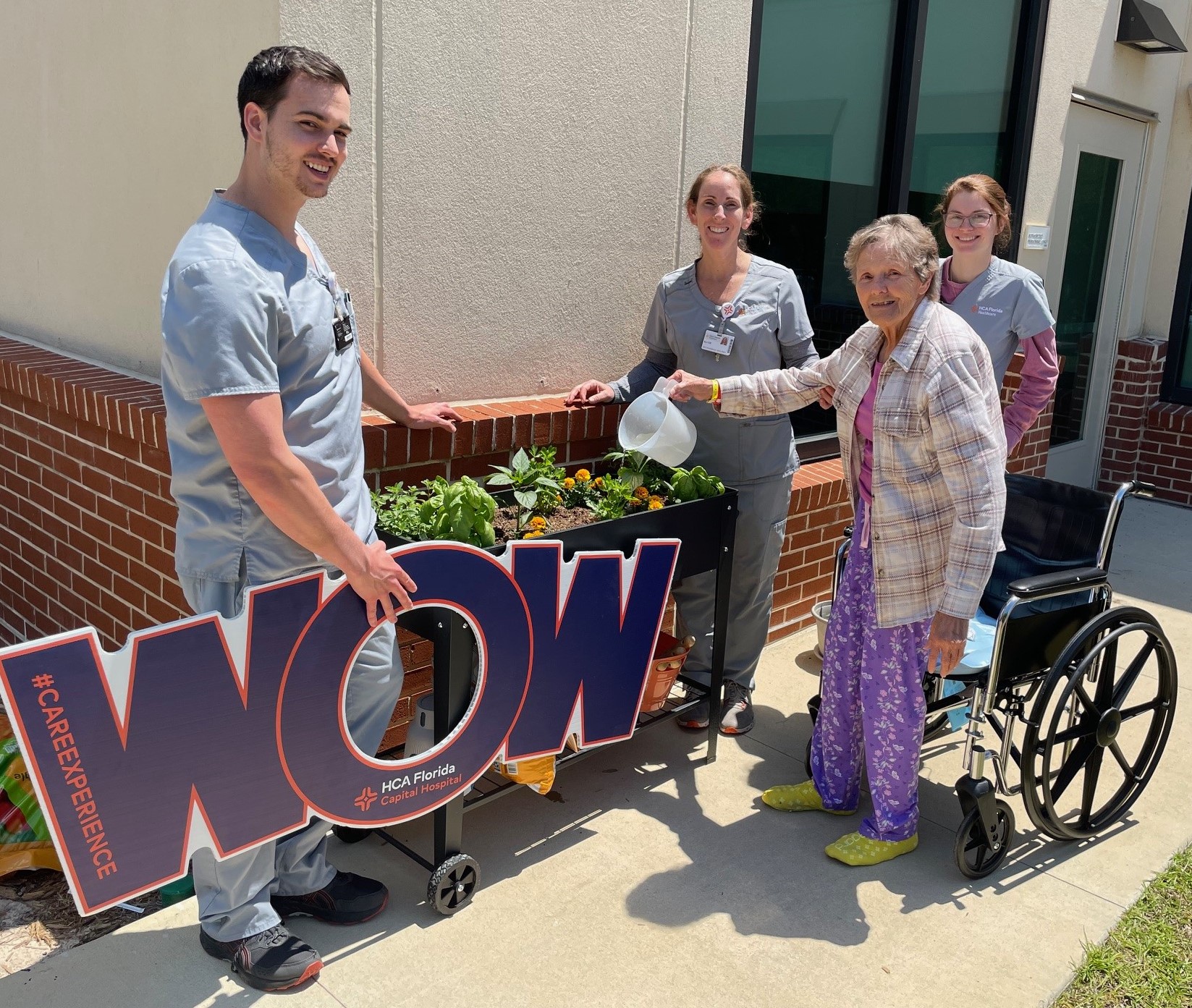 Agnes standing beside wheelchair watering flowers with physicians holding a big "WOW" sign