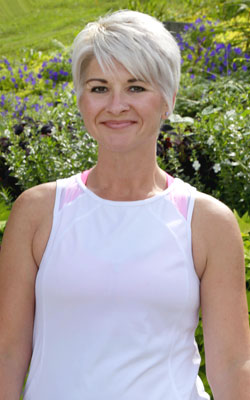 Kendra smiles while wearing a white workout top and is standing in a garden.
