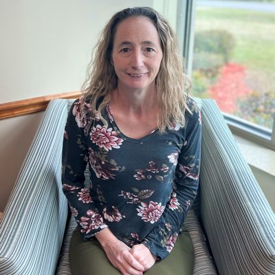 Becky Hammock smiles while wearing a flower print shirt and sitting in a chair at the hospital.