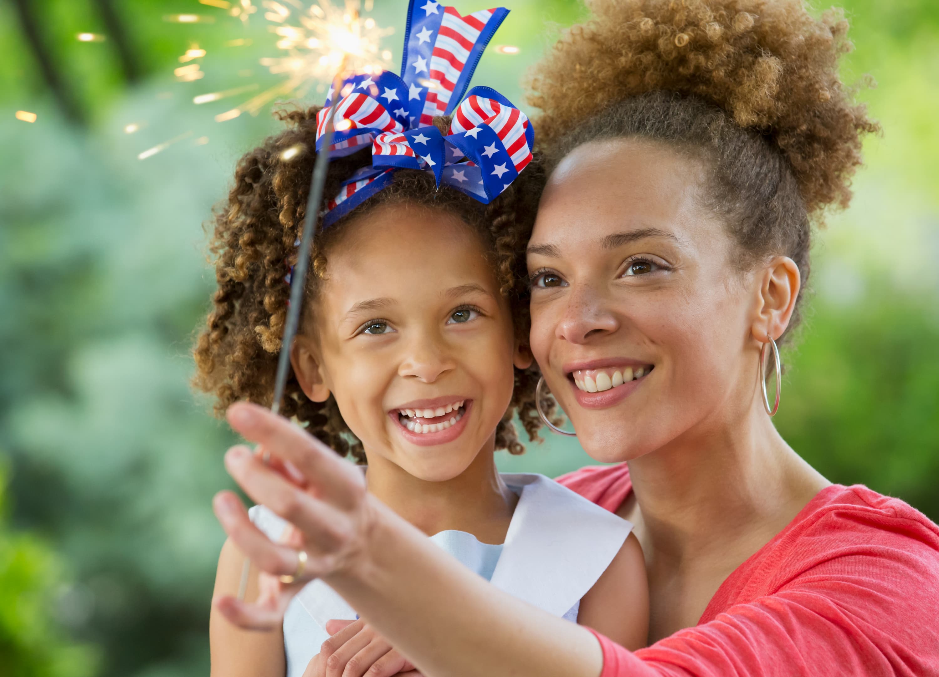 Mother and daughter smiling outdoors while holding a lit sparkler during a summer celebration.