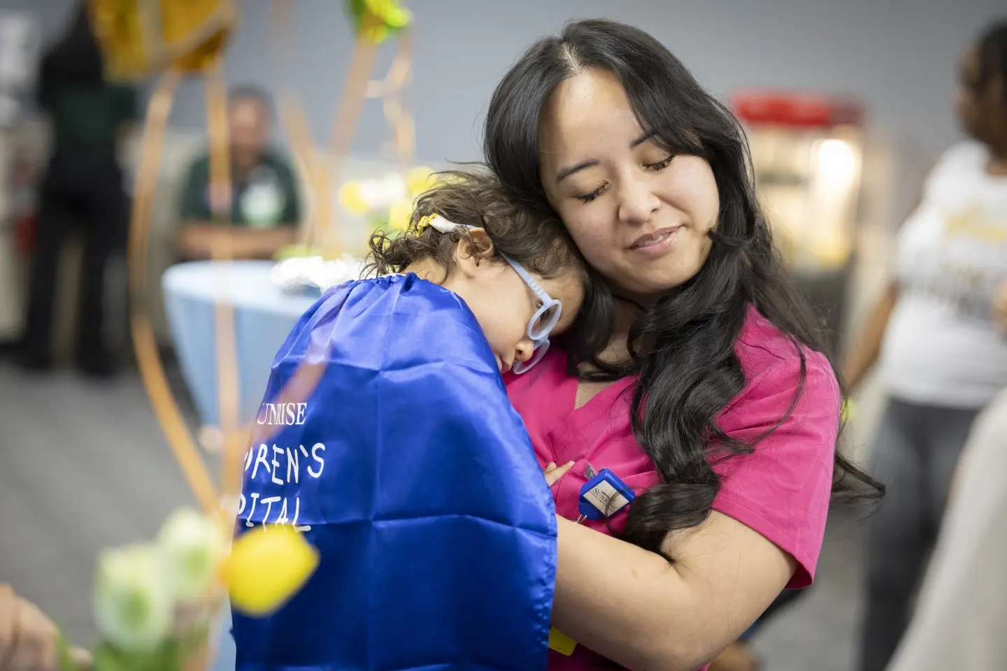 A young NICU nurse with her eyes closed, holding a child wearing a cape and glasses.