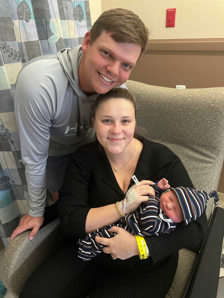 Mother Taylor holds her newborn William while seated in a recliner, with father Luke standing behind them smiling.