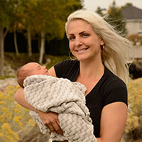 Jessica smiles while holding her newborn in front of their house.