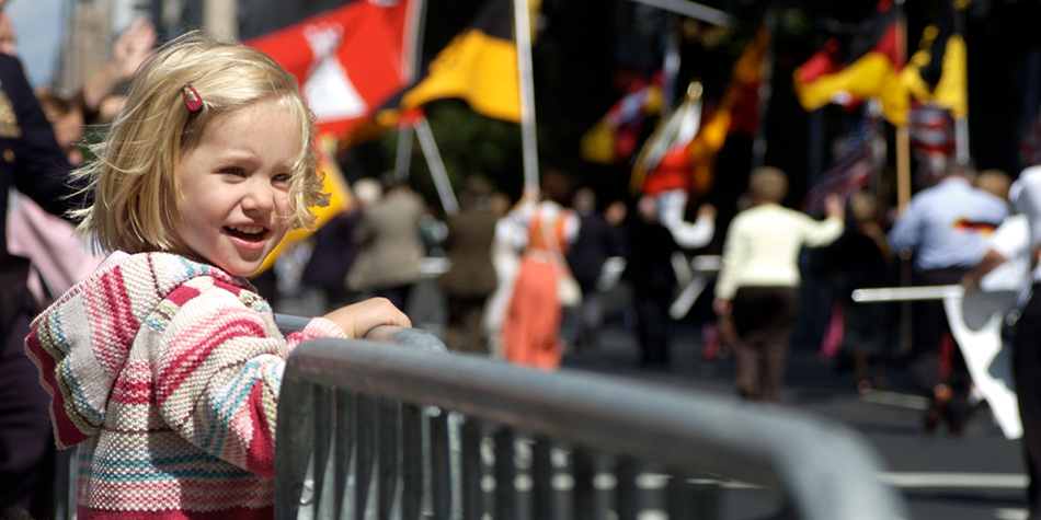 A child smiles while standing on a sidewalk, watching a summertime parade.