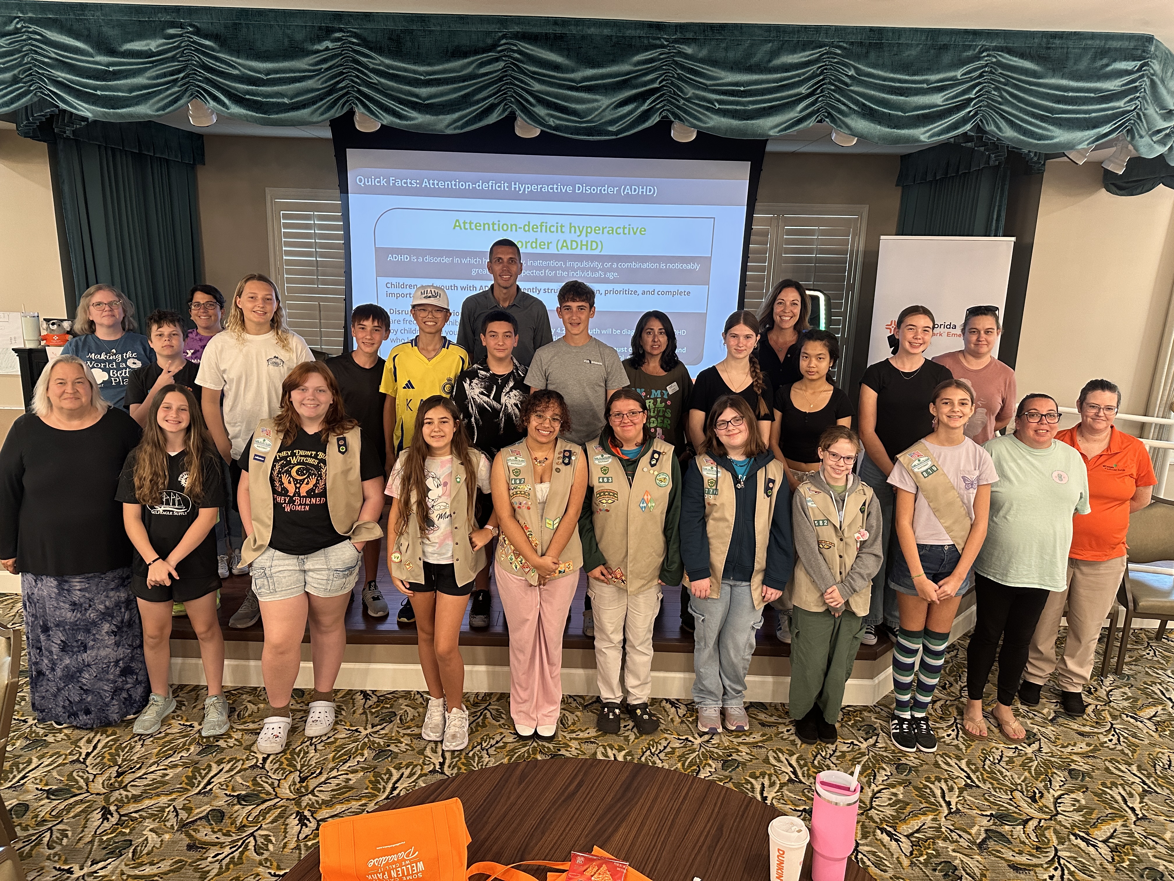 A group of people, including youth and adults, pose together in a room during a Youth Mental Health First Aid training. Some participants wear uniforms with patches and badges. Behind them, a presentation slide on ADHD is displayed. The room features green curtains, patterned carpet, and tables with personal items in the foreground.