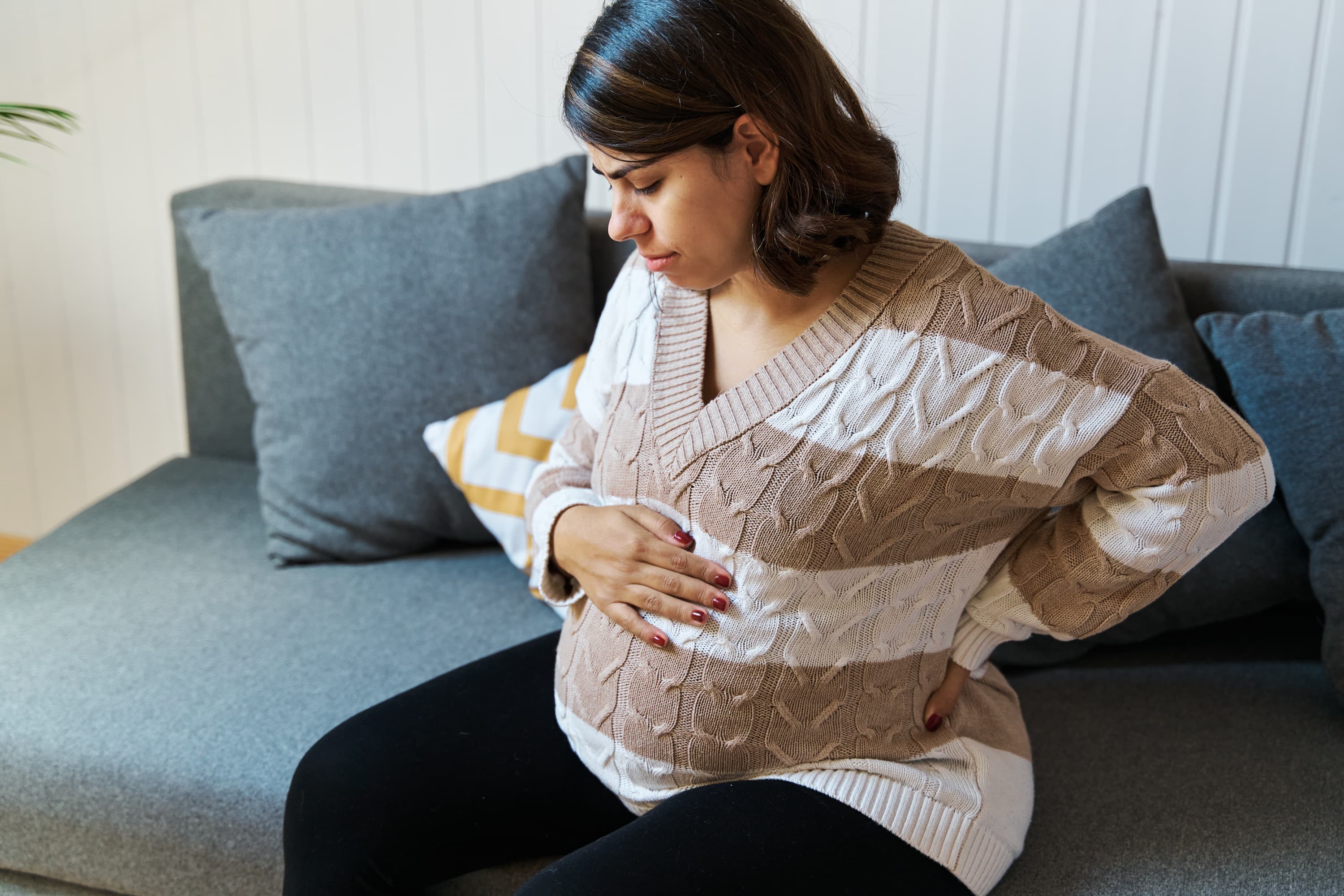 A pregnant woman holds her stomach and holds her back while sitting on a couch.