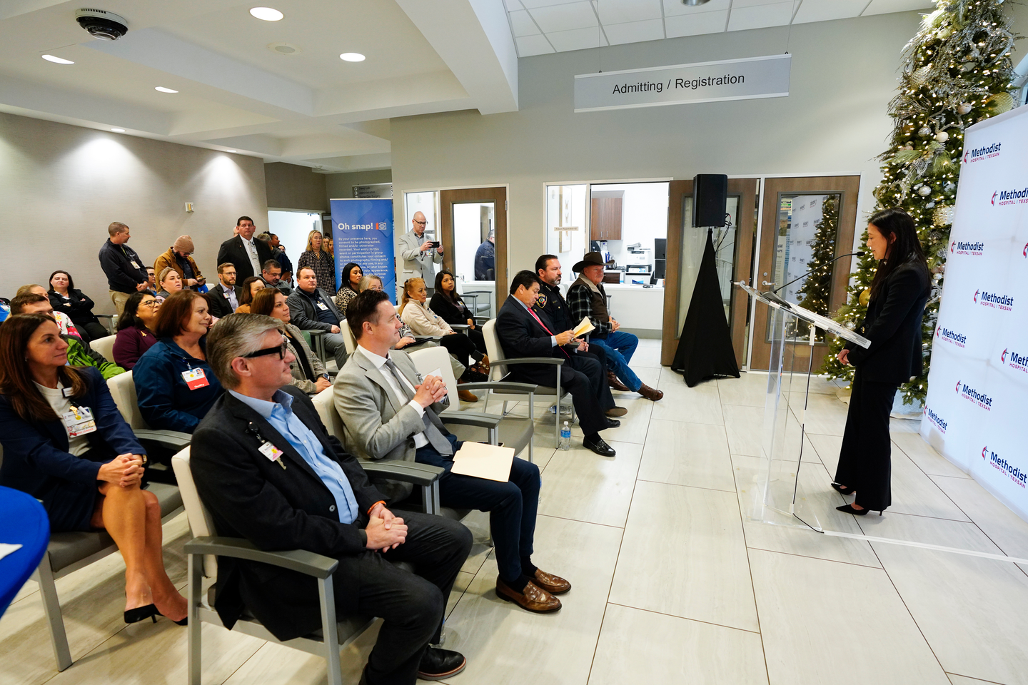A speaker stands at a podium addressing an audience seated in a hospital lobby area. The room features holiday decorations, including a large Christmas tree, and signage for Methodist Hospital Texsan is visible in the background.