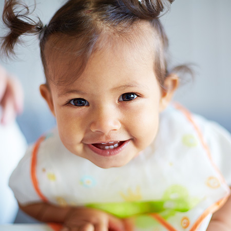 A baby with pigtails and an orange and green bib smiles while lying on their stomach.