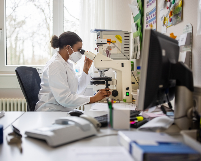 A medical researcher in a lab coat at a desk, looking at a sample in a microscope.