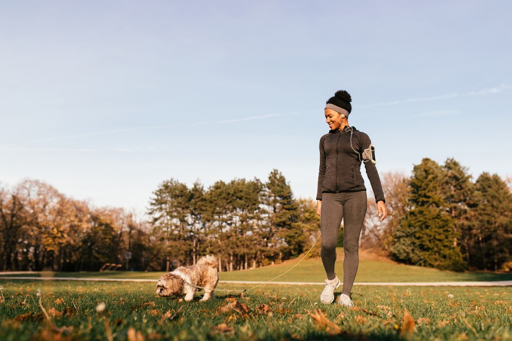 A woman in running gear stands with her dog in a field of grass.