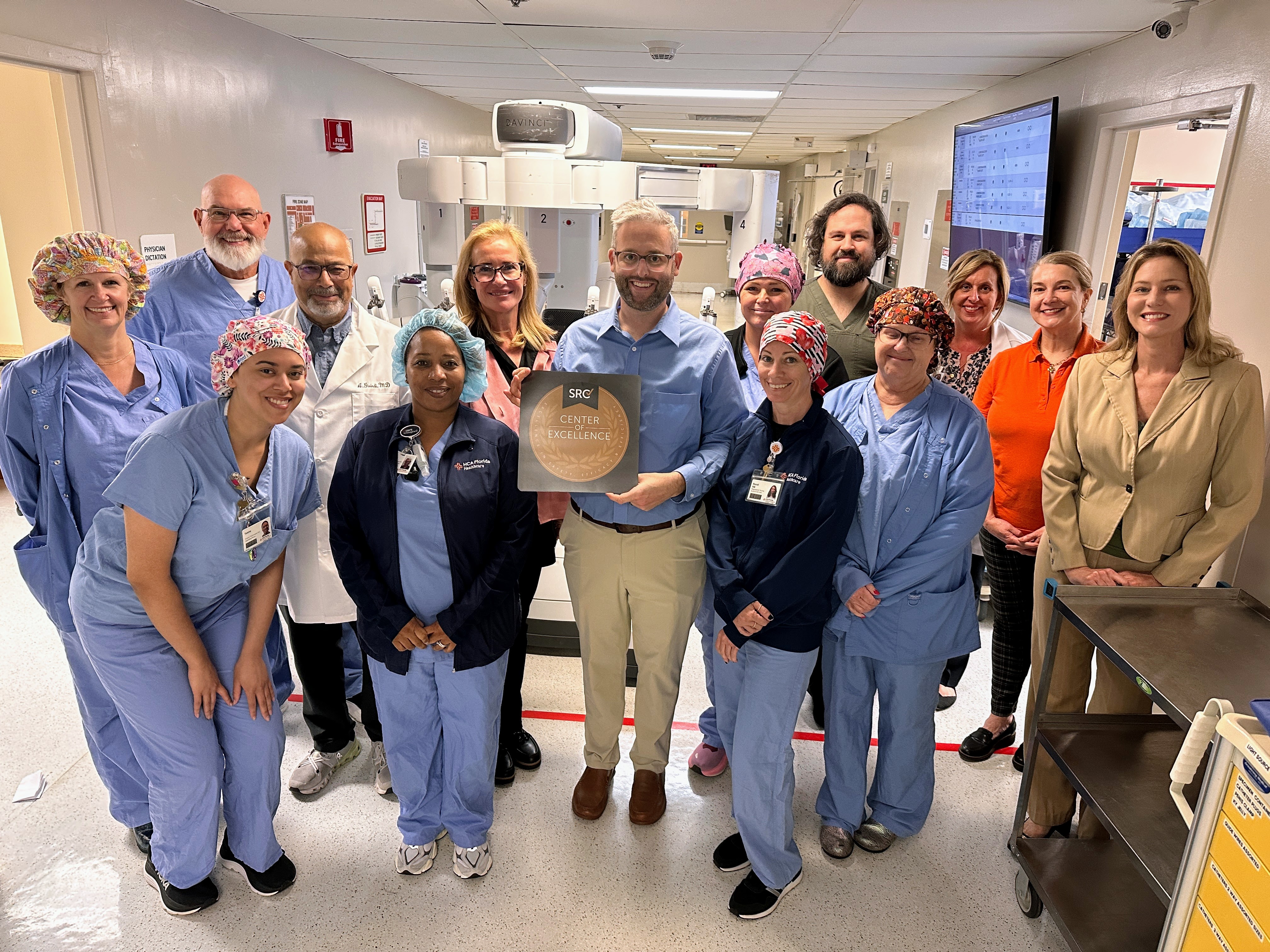 A group of surgeons, nurses and clinical staff at HCA Florida Englewood Hospital stand together in a surgical suite, smiling and holding a Center of Excellence plaque in front of robotic surgery equipment.