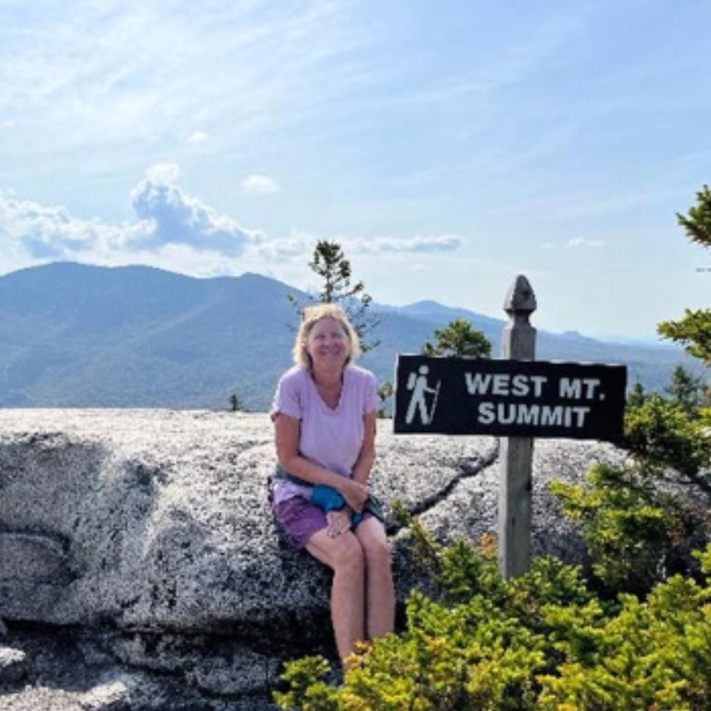 Carolyn sitting on a large rock, next to a sign that reads 'West Mt. Summit' on a hiking trail.