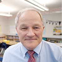 Garry smile while sitting in a school classroom, wearing a pale blue button-up shirt and a red tie.