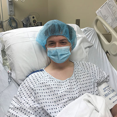 Jarrett Johnson laying in a hospital bed wearing a mask, hospital gown, and blue hair net.