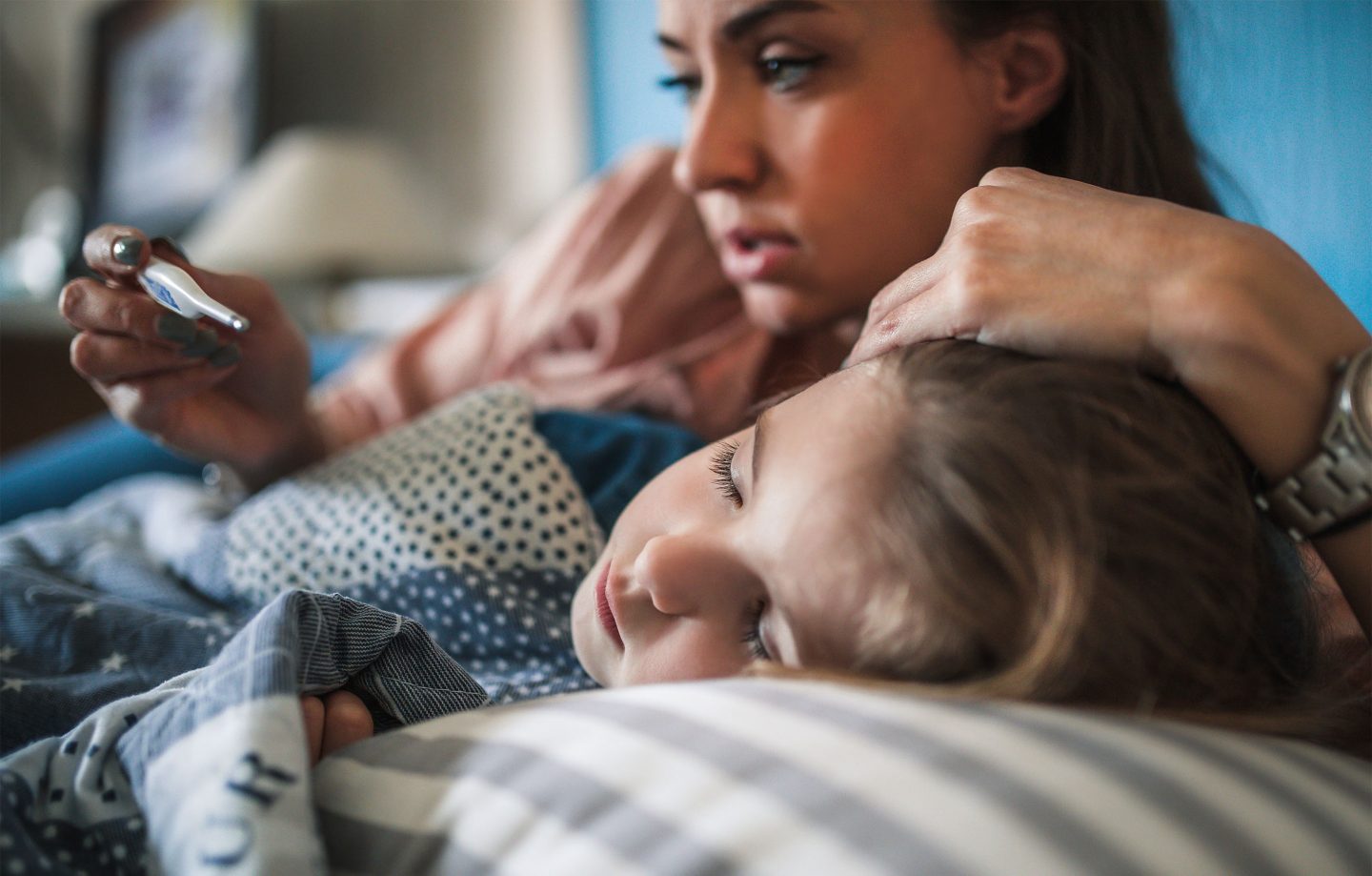 A woman lies in bed with her sick child, looking at a thermometer.
